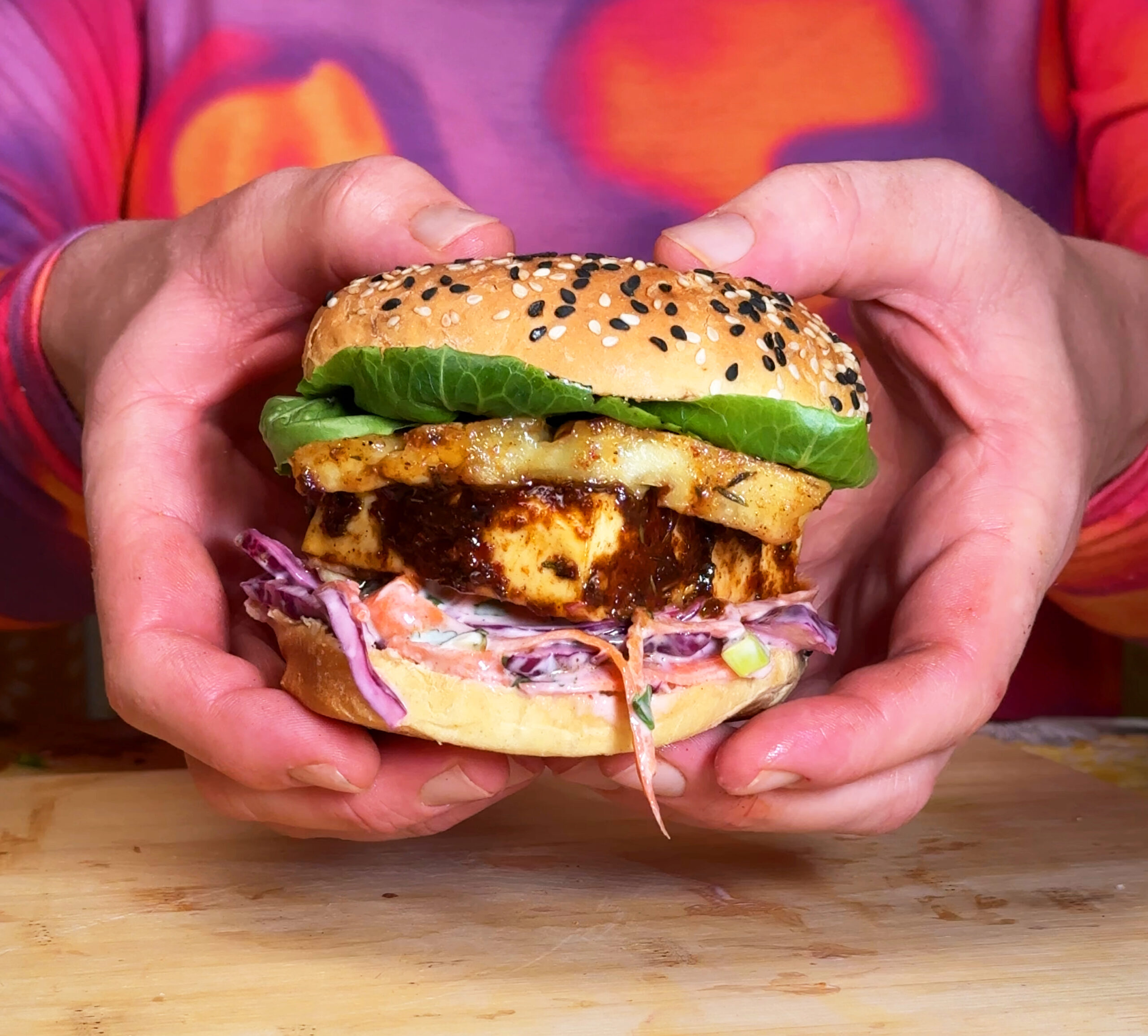 A burger being held in two hands featuring a brioche bun filled with jerk spiced paneer, burnt pineapple, crunchy slaw and lettuce.